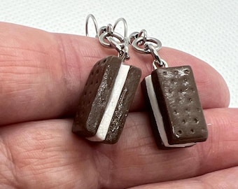 May include: A pair of novelty earrings featuring miniature ice cream sandwiches. The earrings have a dark brown, textured cookie exterior with a white cream filling. They hang from silver-colored ear wires, offering a playful and unique accessory.
