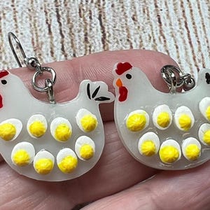 May include: Novelty earrings shaped like white chickens with red combs and yellow egg yolks. The earrings are made of a translucent material and have silver-coloured ear wires.