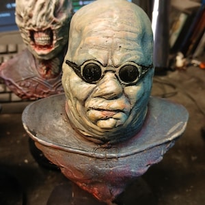 May include: A detailed bust sculpture of a creature with a pale blue-green skin tone, wearing goggles. The creature has a wrinkled forehead and a wide, flat nose. The bust is painted with a variety of colors, including blue, green, red, and brown.
