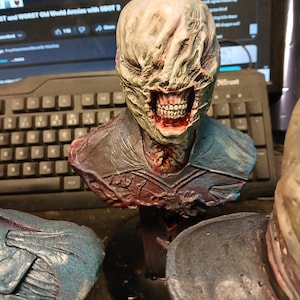 May include: A detailed bust of a horror movie monster with a pale, fleshy face, a wide, toothy grin, and a torn, raggedy suit. The bust is painted in shades of gray, brown, and red.