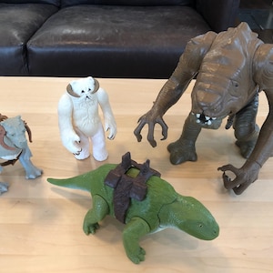 Puede incluir: Una colección de figuras de acción de Star Wars, que incluye un monstruo Rancor marrón, un Wampa blanco, una criatura verde con forma de dinosaurio y otras dos figuras con monturas. Las figuras están dispuestas sobre una superficie de madera clara.