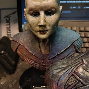 May include: A painted bust sculpture of a person with a bald head and a colorful, textured, fabric garment. The bust is on a black circular base.