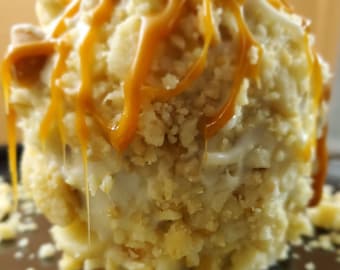 May include: A caramel apple coated in white chocolate and crushed cookies, with caramel drizzle. The dessert is on a stick and sits on a dark surface. The image is a close-up shot of the sweet treat.