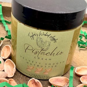 May include: A jar of pistachio cream nut butter with a black lid. The label reads "Taylor's Wicked Apples Pistachio Cream Velvety Smooth Nutbutter." Pistachio shells and green paper ribbon surround the jar.