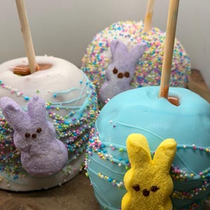 Peut inclure: Trois pommes d'amour sur bâtonnets, chacune avec un enrobage et une décoration différents. Une est blanche avec un filet bleu et un lapin violet, une autre est bleue avec un lapin jaune, et la troisième est recouverte de vermicelles avec un lapin violet.