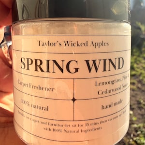 Könnte beinhalten: Ein klares Glas mit schwarzem Deckel, beschriftet mit "Taylor's Wicked Apples SPRING WIND Carpet Freshener". Das Etikett enthält Text über die natürlichen Inhaltsstoffe und Duftnoten von Zitronengras, Kiefer und Zedernholz. Das Glas wird von einer Person gehalten.