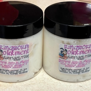 May include: Two jars of ringworm ointment with black lids and white labels. The labels feature text and a cartoon illustration. The ointment is a white, creamy substance. The text on the label includes directions and ingredients.