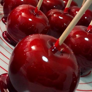 Op de afbeelding: Close-up van meerdere glanzende, rode toffee-appels op houten stokjes. De appels zijn bedekt met een glanzende, donkerrode snoepcoating. De toffee-appels zijn gerangschikt op een wit oppervlak met rode lijnen.