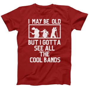 K&ouml;nnte beinhalten: Rotes T-Shirt mit wei&szlig;em Text, der "I may be old but I gotta see all the cool bands" lautet. Der Text ist von einem wei&szlig;en Rechteck umgeben. Im Rechteck befindet sich eine Silhouette einer Band, die Musik spielt.
