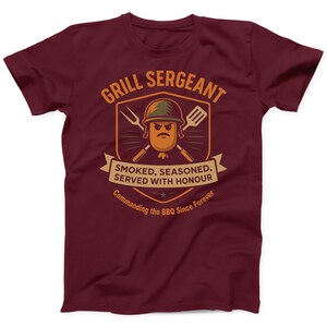 May include: Burgundy t-shirt with a humorous graphic. The design features a cartoon sausage wearing a military helmet, crossed grilling tools, and the text "GRILL SERGEANT". Below, a banner reads "SMOKED, SEASONED, SERVED WITH HONOUR" and "Commanding the BBQ Since Forever".