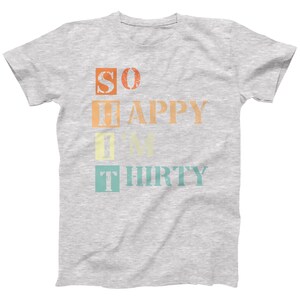May include: A grey t-shirt with the text "SO HAPPY I'M THIRTY" printed in colourful boxes. The text is in a vintage style font.