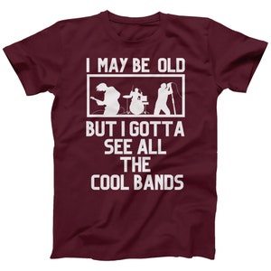 K&ouml;nnte beinhalten: Burgunderfarbenes T-Shirt mit wei&szlig;em Text, der "I may be old but I gotta see all the cool bands" lautet. Der Text ist von einem wei&szlig;en Rechteck umgeben. Im Inneren des Rechtecks befindet sich eine Silhouette einer Band, die Musik spielt.