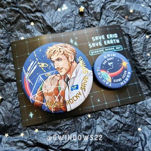 May include: Two circular pin-back buttons featuring space-themed illustrations. The larger button depicts a blonde person holding a rock, with text reading 'GRACE ROCKY SAVE STARS'. The smaller button shows a planet and text 'ERID HAIL MARY TAU CETI ROCKY GRACE EARTH'.