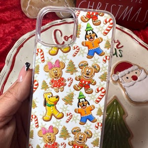 May include: Clear phone case featuring a festive pattern of gingerbread characters, Christmas trees, and candy canes. The case showcases Mickey and Minnie Mouse, Pluto, and Goofy in gingerbread form, set against a white background. A holiday-themed accessory.