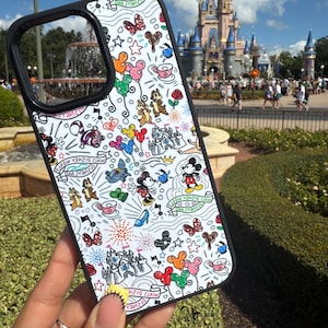 May include: A white phone case with a black border, featuring a whimsical Disney-themed design. The case is adorned with colorful illustrations of Mickey Mouse, Minnie Mouse, Chip and Dale, castles, and other iconic elements. The design includes the text "Walt Disney World".