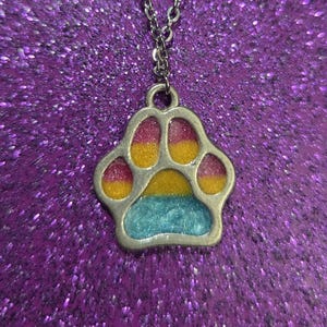 May include: A silver-toned necklace with a paw print pendant. The paw print is filled with pink, yellow, and blue enamel. The chain has small, round beads near the clasp. The background is a sparkly, purple surface.