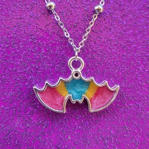 May include: A silver-colored necklace with a bat pendant. The bat pendant has a silver outline and is filled with pink, yellow, and teal colors. The necklace chain is delicate and has small silver beads.