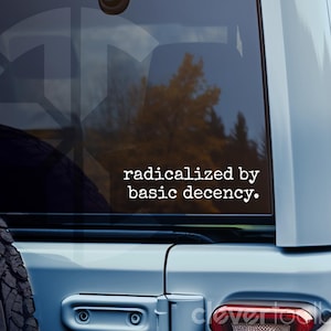 Könnte beinhalten: Ein weißer Vinyl-Aufkleber auf der Heckscheibe eines hellblauen Fahrzeugs mit dem Text "radicalized by basic decency." in einer Schreibmaschinen-Schriftart. Auf der linken Seite ist ein Ersatzrad am Fahrzeug montiert.