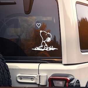 May include: A white vinyl decal applied to a vehicle window featuring a line art illustration of a cartoon dog sitting in grass and looking up at a small heart shape. The design is rendered in simple white outlines against the dark glass surface.