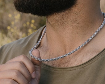 May include: A silver-colored rope chain necklace. The necklace is worn around the neck and is made of tightly twisted metal links. The person in the image is wearing a green shirt.