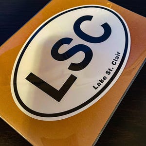 Puede incluir: Una pegatina ovalada con las letras "LSC" en negrita negra sobre un fondo blanco. Las palabras "Lake St. Clair" están impresas en negro a lo largo del borde inferior de la pegatina. La pegatina está sobre una superficie marrón.