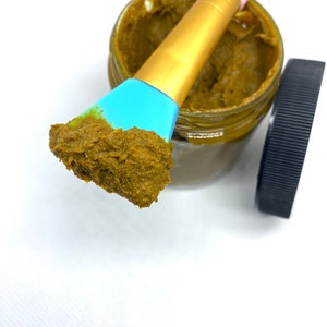 Pu&ograve; includere: Un piccolo barattolo di vetro pieno di una maschera per il viso marrone, verde e gialla. Un pennello blu e dorato &egrave; immerso nel barattolo e ha una piccola quantit&agrave; di maschera su di esso.