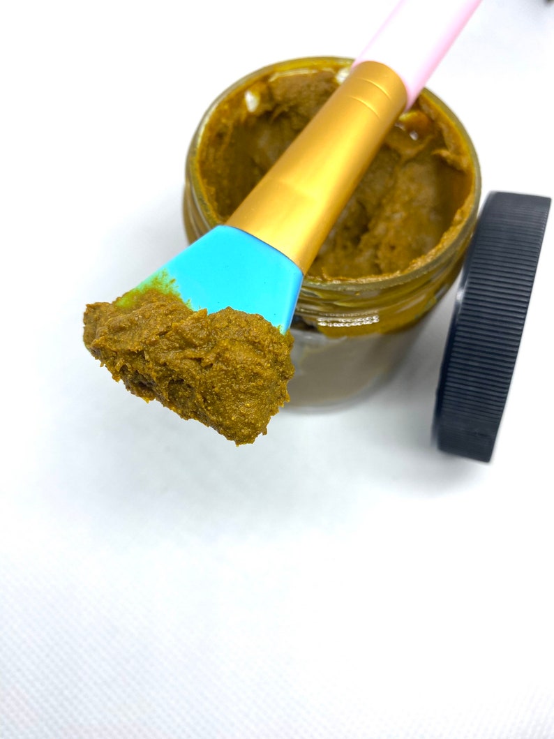 Pu&ograve; includere: Un piccolo barattolo di vetro pieno di una maschera per il viso marrone, verde e gialla. Un pennello blu e dorato &egrave; immerso nel barattolo e ha una piccola quantit&agrave; di maschera su di esso.