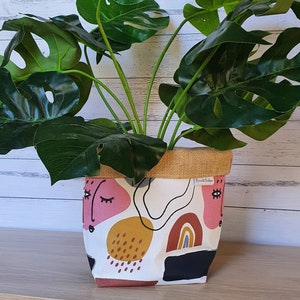 May include: A white fabric planter with a hessian top and a colourful abstract design featuring pink, yellow, brown, and black shapes. The planter is filled with a green monstera plant.