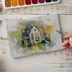 May include: A watercolor painting of a house with a green lawn and a blue sky. The painting is in a sketchbook with a pencil and eraser nearby. There are also watercolor paints and pencils on the table.