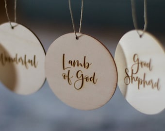 May include: Four round wooden ornaments with twine hangers. Each ornament has a different phrase written in cursive: "Wonderful", "Lamb of God", "Good Shepherd", and "Light is".