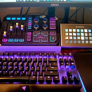 May include: A black keyboard with purple backlighting, a black and blue audio mixer with a screen that says "GoX", and a black Stream Deck with a gray face and a grid of buttons.