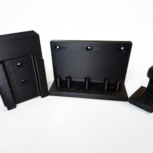 May include: Three black 3D-printed wall mount brackets. The brackets are rectangular with mounting holes and various shapes for holding items. The brackets are made of a matte black material and are displayed against a white background.