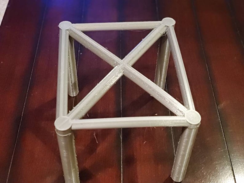 May include: A white, 3D printed, square-shaped stand with four legs and a criss-cross design.