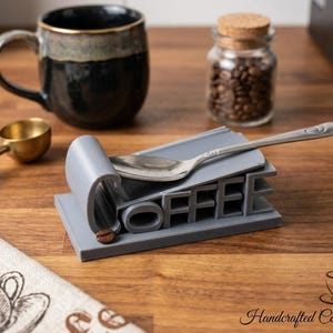 May include: A gray coffee spoon rest with the word "COFFEE" cut out of the design. A silver spoon rests on top. A coffee bean sits in the "O". A coffee cup, coffee beans, and a measuring spoon are also visible.