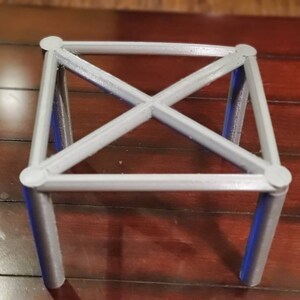 May include: A gray, 3D printed, square-shaped stand with four legs and a criss-cross design.