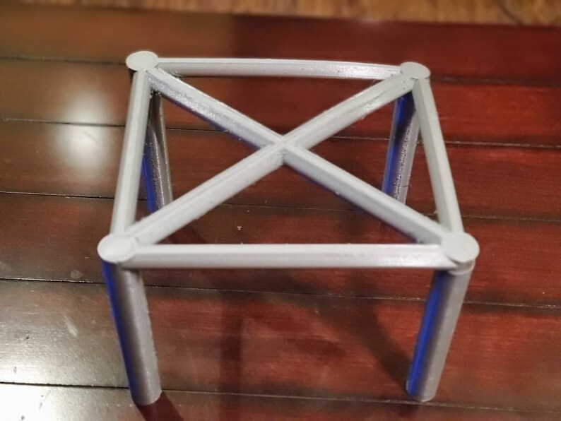 May include: A gray, 3D printed, square-shaped stand with four legs and a criss-cross design.