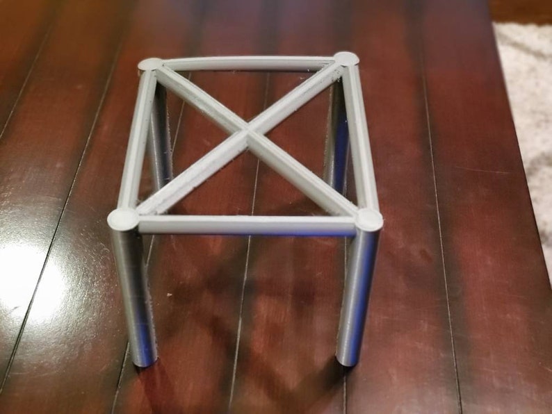 May include: A white metal stand with four legs and a criss-cross design. The stand is made of metal tubing and has a square base.