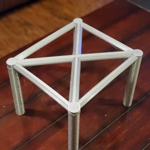 May include: A white, geometric, metal stand with four legs and a square top. The stand is made of a lightweight material and has a simple, modern design.