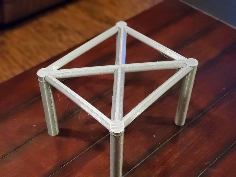 May include: A white, geometric, metal stand with four legs and a square top. The stand is made of a lightweight material and has a simple, modern design.