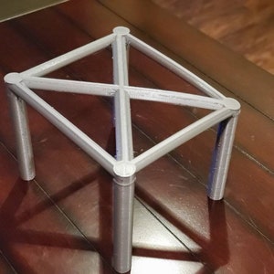 May include: A silver metal stand with four legs and a diamond-shaped top. The stand is made of thin metal rods that are connected at the corners and in the center.