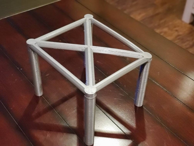 May include: A silver metal stand with four legs and a diamond-shaped top. The stand is made of thin metal rods that are connected at the corners and in the center.