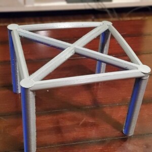 May include: A silver metal stand with four legs and a square top. The stand is made of thin metal rods that are connected at the corners and in the center.