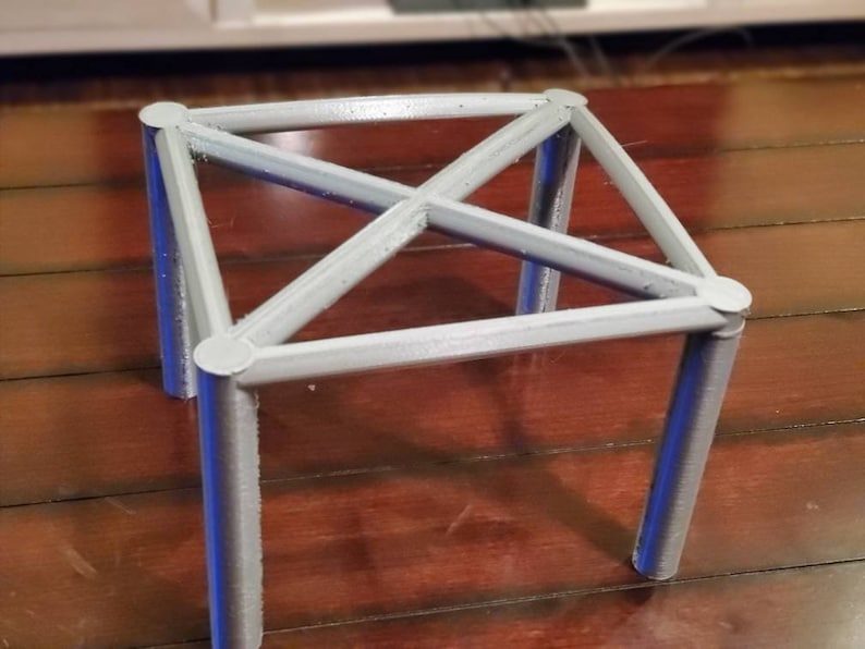May include: A silver metal stand with four legs and a square top. The stand is made of thin metal rods that are connected at the corners and in the center.