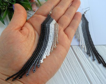 May include: A pair of long, beaded earrings with a graduated color scheme of black, white, and silver. The earrings are hanging from a person's hand.