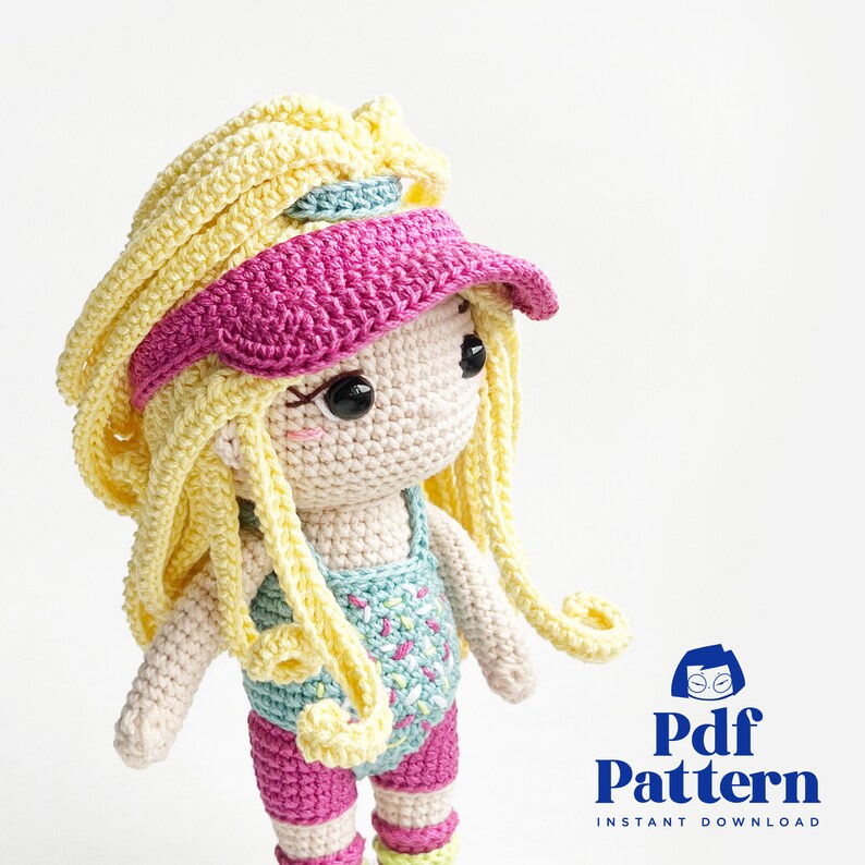 May include: Crochet doll pattern for a girl with long yellow hair, wearing a pink visor, a blue and white swimsuit, and pink shorts. The doll is holding a pink and white striped ball. The pattern is available as an instant download.