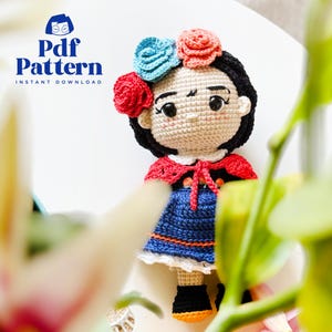 以下が含まれることがあります： 花飾りのついたかぎ針編みの人形、赤いショール、青いドレス。人形は黒髪で、オレンジ色のソールが付いた黒い靴を履いています。画像には「Pdf Pattern Instant Download」というテキストが含まれています。