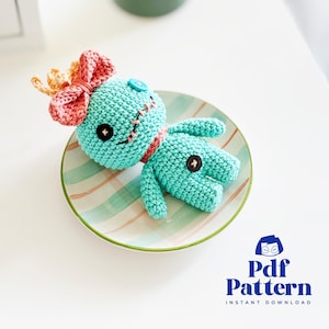 Puede incluir: Una muñeca azul de crochet con un lazo rosa y pelo amarillo. La muñeca tiene una nariz de botón y una boca cosida. La muñeca está tumbada en un plato rayado.