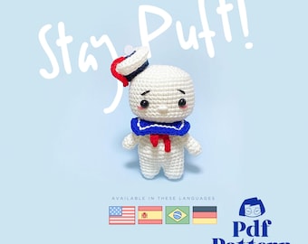 May include: A crocheted Stay Puft Marshmallow Man amigurumi doll wearing a blue and red sailor hat. The doll is white with black eyes and a red mouth. The text "Stay Puft!" is written in white above the doll. The text "Pdf Pattern Instant Download" is written below the doll. The text "Available in these languages" is written above four flags: American, Chinese, Brazilian, and German.