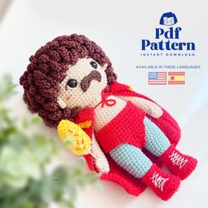 Puede incluir: Una muñeca de ganchillo con pelo castaño rizado, bigote y mejillas sonrosadas. La muñeca lleva una capa roja, pantalones cortos rojos, mallas azul claro y botas rojas con cordones blancos. Sostiene una mazorca de maíz amarilla. Texto: Pdf Pattern.