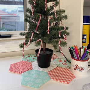Può includere: Un piccolo albero di Natale artificiale decorato con bastoncini di zucchero a righe rosse e bianche. L'albero è in un vaso nero su una superficie bianca, circondato da un sottobicchiere in tessuto colorato a fantasia. È visibile una tazza con la scritta "Joy".